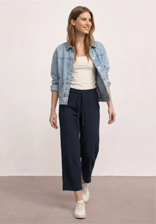 Frau in weiter, dunkler Hose, weißem Top und Jeansjacke, trägt weiße Sneaker vor neutralem Hintergrund.