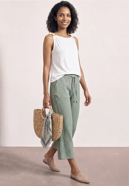 Vrouw in witte top, groene 7/8-broek en rieten tas met sjaal voor neutrale achtergrond.