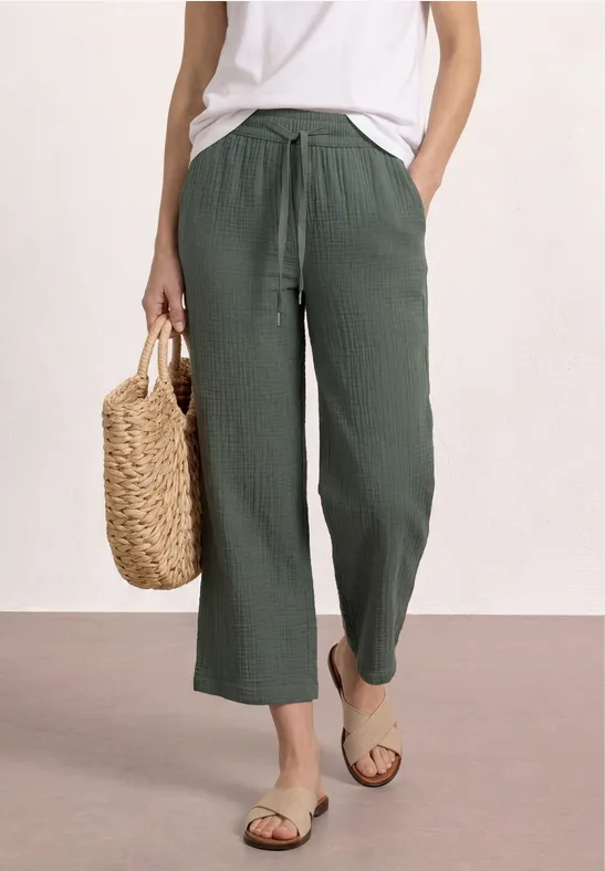 Vrouw in een wit topje en groene linnen broek, met strozak en sandalen.