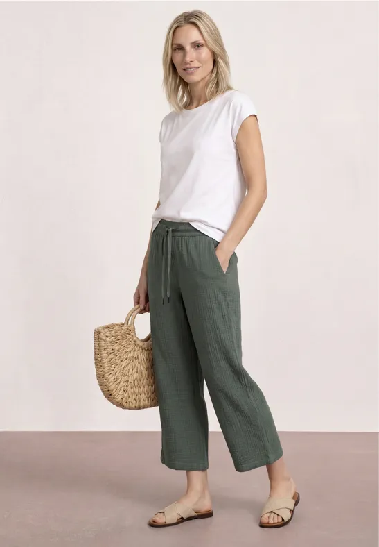 Vrouw in wit T-shirt, groene wijde broek, sandalen en strooien tas tegen lichte achtergrond.