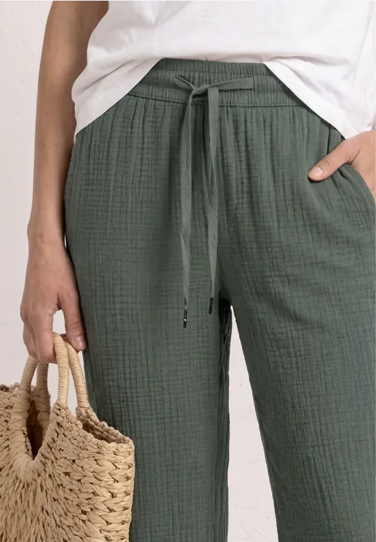 Persoon draagt wit shirt en groene, losse broek met trekkoord en houdt een gevlochten strozak vast.