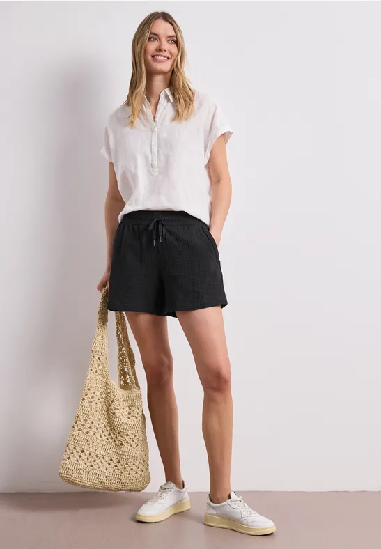 Junge Frau in weißer Bluse, schwarzen Shorts, Sneakers hält eine geflochtene Tasche.