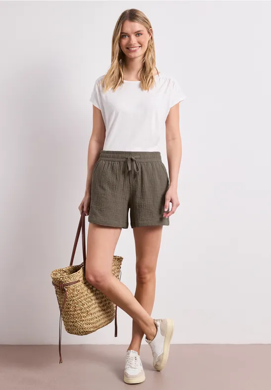 Femme en t-shirt blanc et short vert olive, tenant un panier tressé, chaussée de baskets blanches.