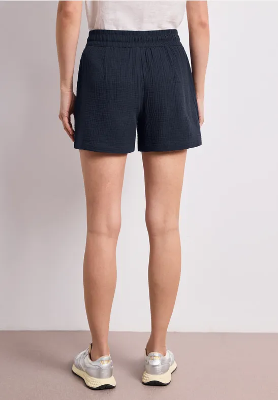 Rückansicht einer Person mit dunkelblauen Shorts, weißem Oberteil und Sneakern vor hellem Hintergrund.