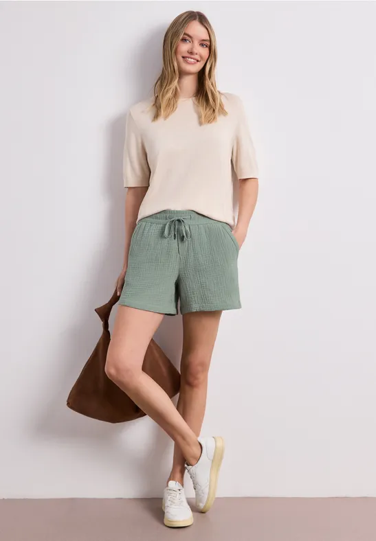 Frau in beiger Bluse, grünen Leinen-Shorts, weißen Sneakern und brauner Tasche vor schlichtem Hintergrund.
