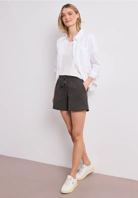 Eine Frau steht, trägt weiße Bluse, schwarze Shorts und weiße Sneaker.