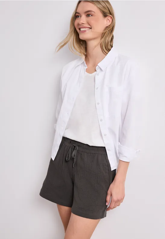 Frau in weißem Hemd und schwarzen Shorts mit Kordelzug vor neutralem Hintergrund.