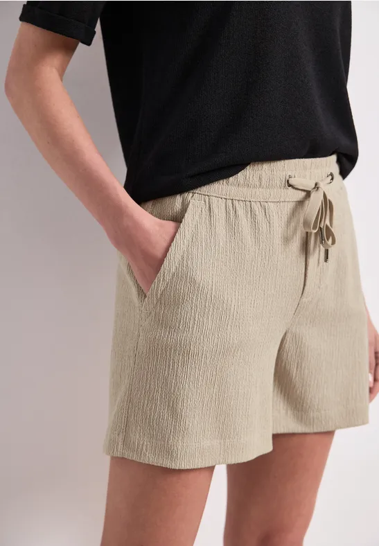 Person trägt beige Shorts mit Tunnelzug, schwarzen T-Shirt, Hand in der Tasche.
