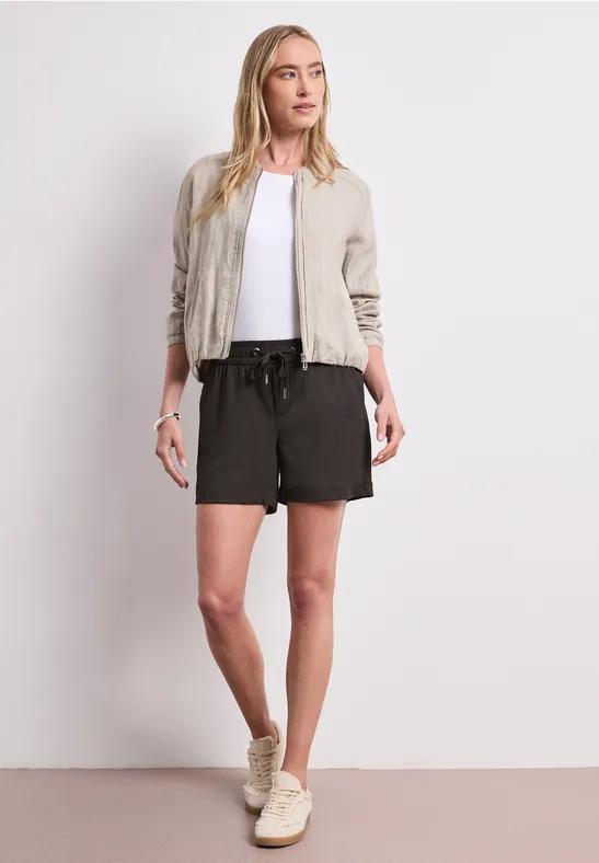 Frau in legerem Outfit mit hellbeiger Jacke, weißem Shirt, schwarzen Shorts und Sneakers.