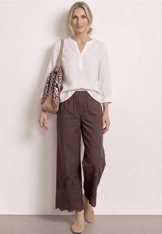 Personne en blouse blanche, pantalon culotte marron à dentelle, chaussures beiges et sac à bandoulière fleuri.