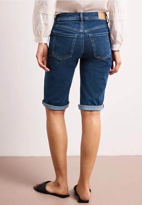 Achteraanzicht van een persoon in knielange opgerolde denimshorts en zwarte instapschoenen.