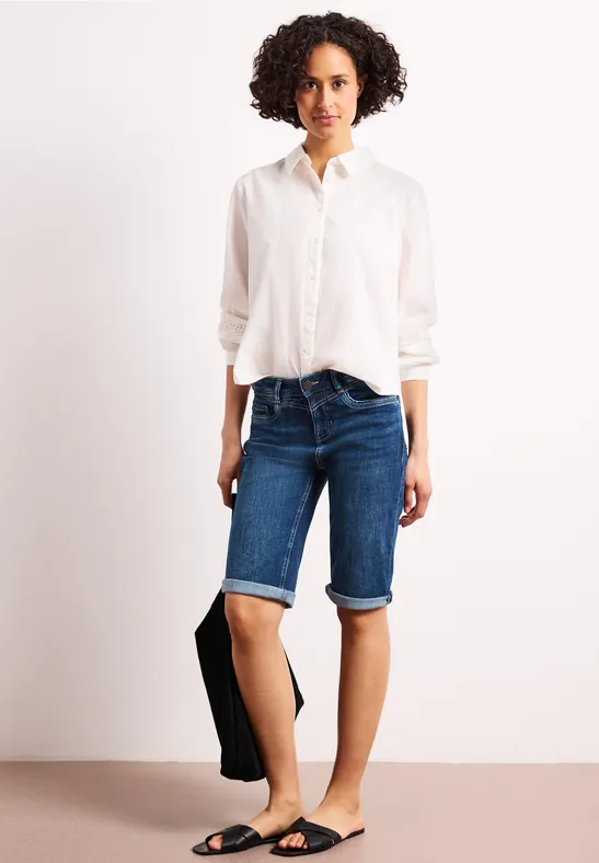 Model draagt wit overhemd, blauwe jeansbermudas, zwarte sandalen en houdt een tas vast.