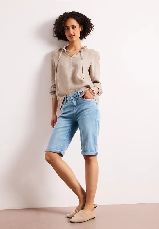 Vrouw in beige linnen blouse en lichtblauwe jeans kniebroek staat voor een witte muur met beige ballerina’s.