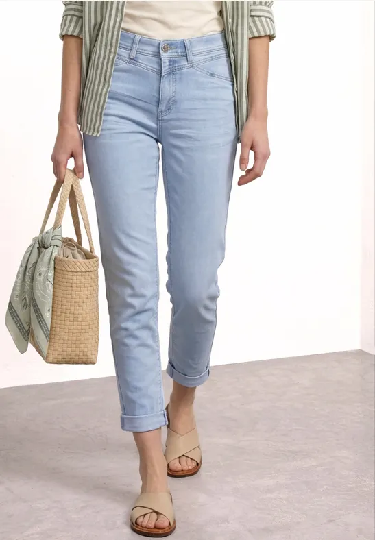 Vrouw in lichtblauwe spijkerbroek en sandalen met een rieten tas met sjaal.