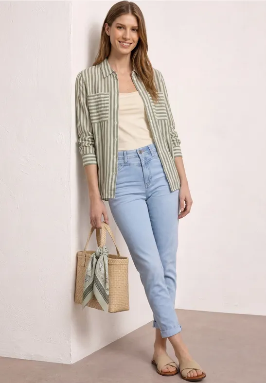 Vrouw voor witte muur, groene gestreepte blouse, top, lichtblauwe jeans, sandalen en strooitas.