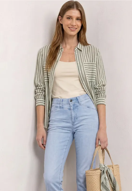 Een vrouw in een wit topje, gestreept overhemd, lichtblauwe jeans en een rieten tas voor een neutrale achtergrond.