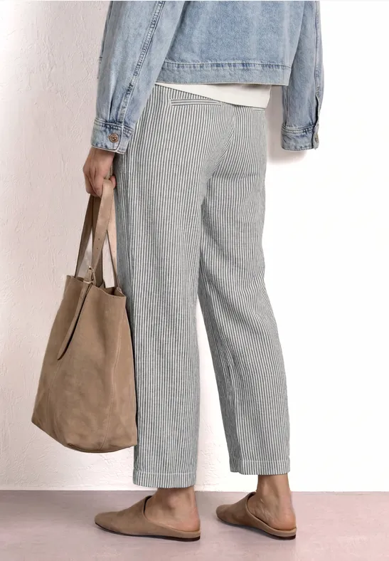 Persoon van opzij met grijze broek, spijkerjas, bruine instappers en een beige suède tas.