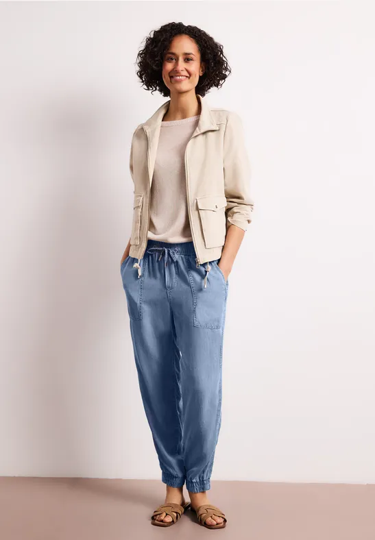 Vrouw in beige blazer, lichte trui, blauwe joggingsbroek en sandalen, handen in de zakken.