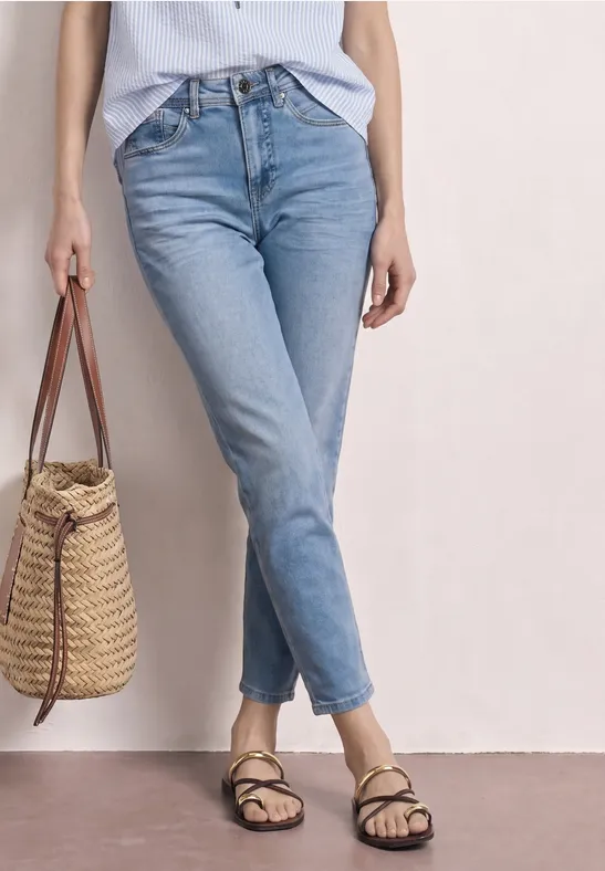 Person trägt hellblaue Jeans, helles Top, schwarze Riemchensandalen und eine geflochtene Strohtasche.