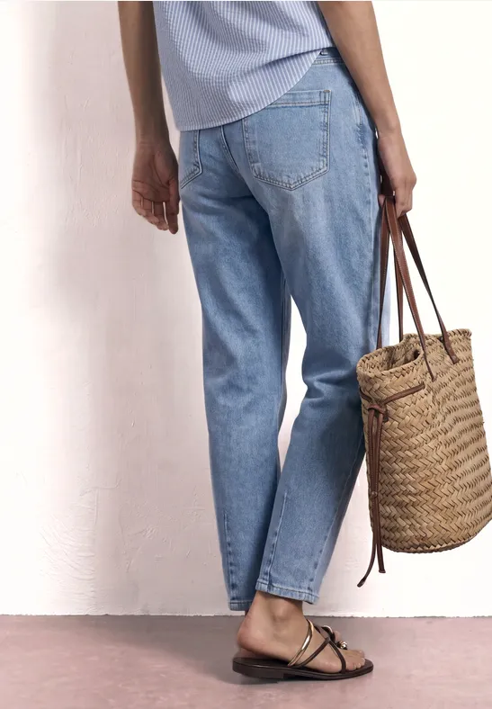 Eine Person in hellen Jeans und Sandalen hält eine geflochtene Strohhandtasche vor einer weißen Wand.