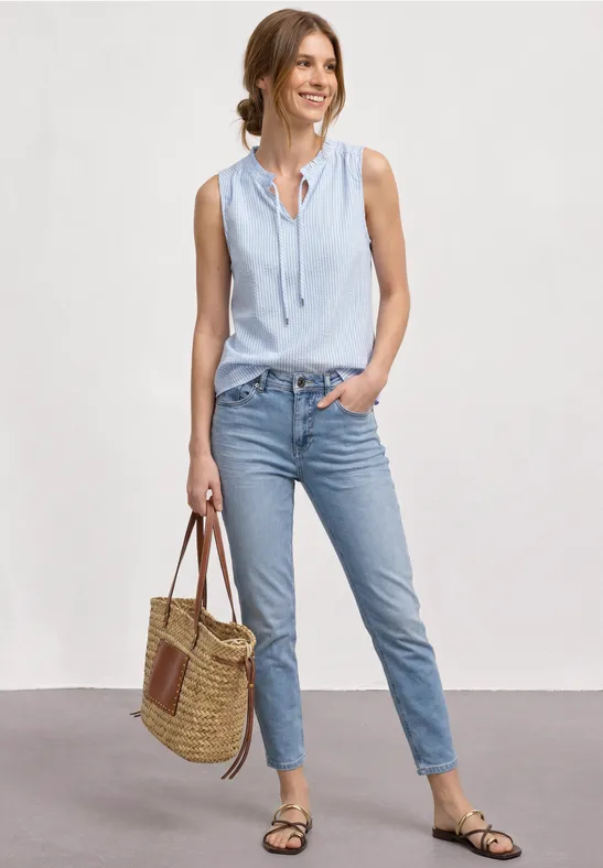 Frau in ärmelloser hellblauer Bluse, hellblauen Jeans, flachen Sandalen und Korbtasche.