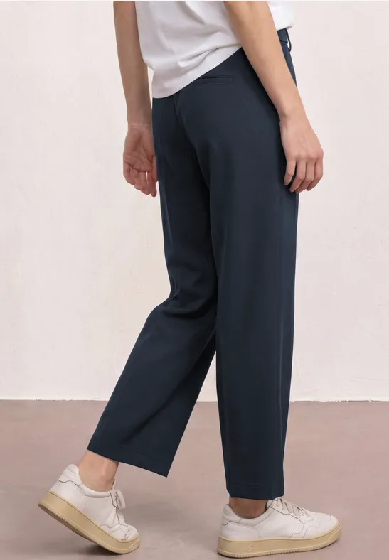 Weibliche Person trägt dunkelblaue, weite 7/8-Hose, weißes Shirt und Sneaker vor hellem, schlichter Kulisse.