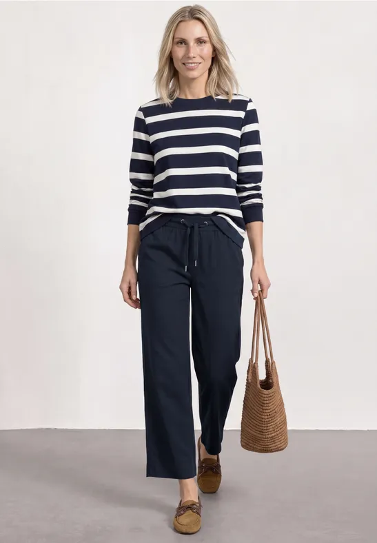 Frau mit blau-weiß gestreiftem Shirt, weiten dunklen Hosen, Basttasche und Loafers.