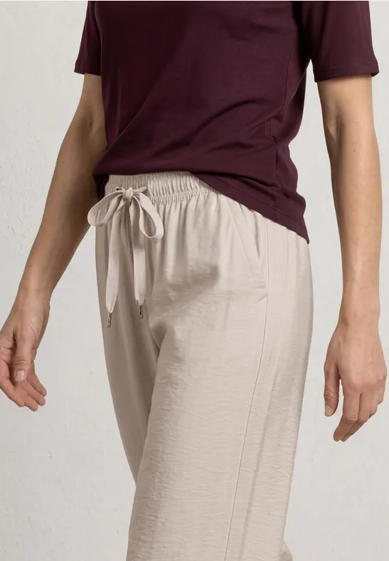 Person trägt helle Leinenhose mit Tunnelzug und dunkelrotes Shirt, vor schlichtem, hellem Hintergrund.