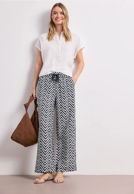 Frau in weißer Bluse und schwarz-weißer Zickzack-Hose mit Schultertasche und Sandalen.