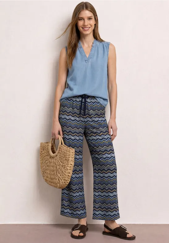 Frau in sommerlichem Outfit: hellblaues ärmelloses Top, gemusterte Palazzohose und Strohtasche, schwarze Sandalen.