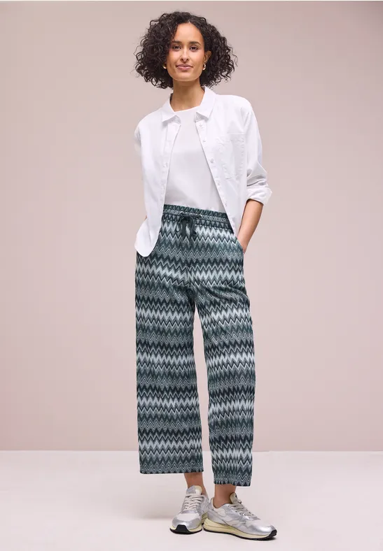 Persoon voor lichte achtergrond, witte blouse op t-shirt, wijde broek met zigzagmotief en metallic sneakers.