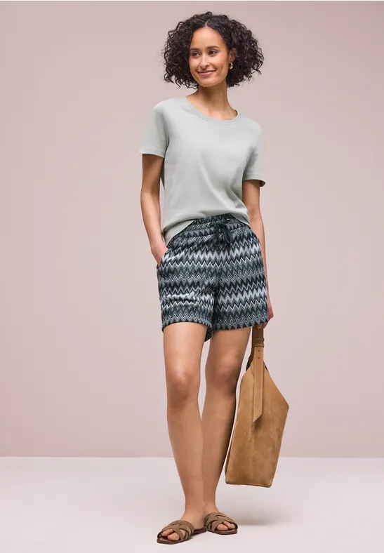 Frau in leichtem T-Shirt, gemusterten Shorts, flachen Sandalen und brauner Tragetasche vor schlichtem Hintergrund.