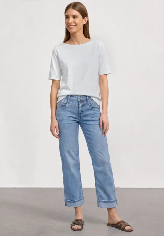 Frau trägt weißes T-Shirt, hellblaue, hochgekrempelte Jeans und braune Sandalen, steht vor neutralem Hintergrund.