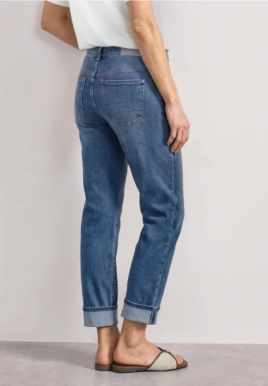 Rückansicht einer Person in blauen, umgeschlagenen Straight-Leg-Jeans und flachen Sandalen, dazu weißes T-Shirt.