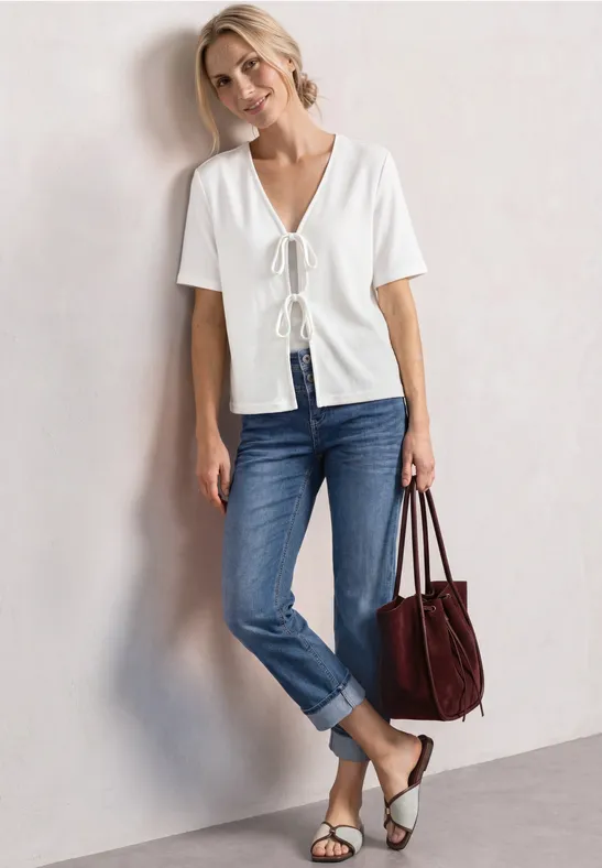 Frau lehnt an der Wand in weißem, vorne gebundenem Shirt, blauem Jeans, Sandalen und roter Tasche.