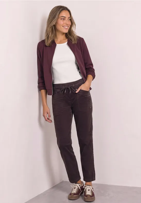 Eine Frau steht in einer Ecke, trägt ein weißes Shirt, bordeauxfarbenen Blazer, dunkle Hose und Sneakers.