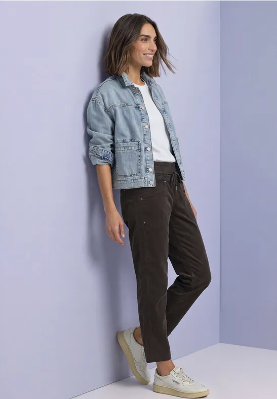 Eine lächelnde Frau in Jeansjacke, weißem Shirt und Cordhosen lehnt an einer pastellfarbenen Wand.