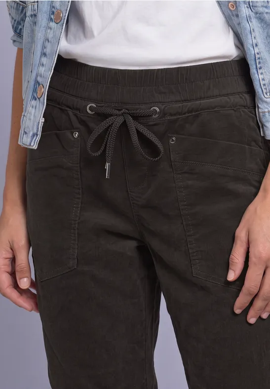 Nahaufnahme einer Person mit dunkelgrüner Cordhose, Tunnelzug und Jeansweste über weißem T-Shirt.