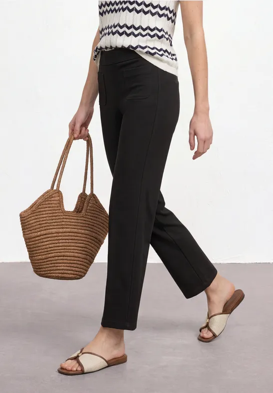 Frau in gemustertem Top, schwarzer Hose, flachen Sandalen und korbförmiger Handtasche