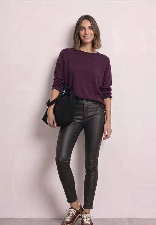 Eine Frau trägt einen lila Pullover, schwarze Lederleggings, Sneakers und hält eine schwarze Tasche vor hellem Hintergrund.