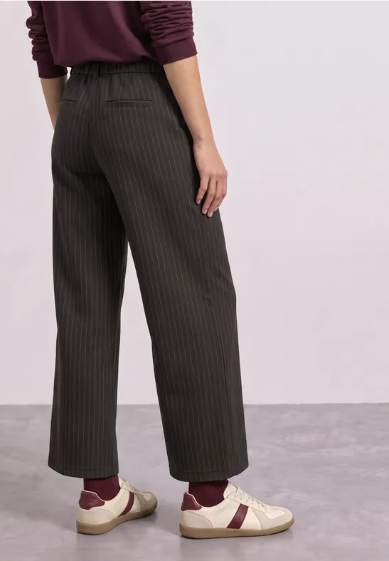Frau trägt schwarze Culottes mit Nadelstreifen, bordeauxfarbene Socken und weiße Slipper auf grauem Boden.