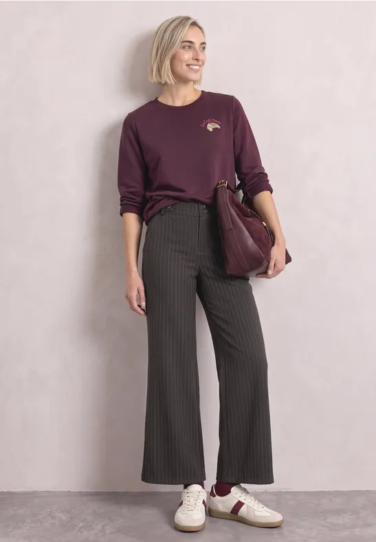 Frau in legerem Chic: burgunderfarbenes Shirt, gestreifte schwarze Schlaghose, weiße Sneaker, bordeauxfarbene Tasche.