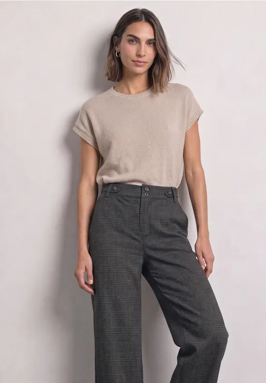Frau trägt beiges T-Shirt und weite dunkelgraue High-Waist-Hosen vor hellem Hintergrund.