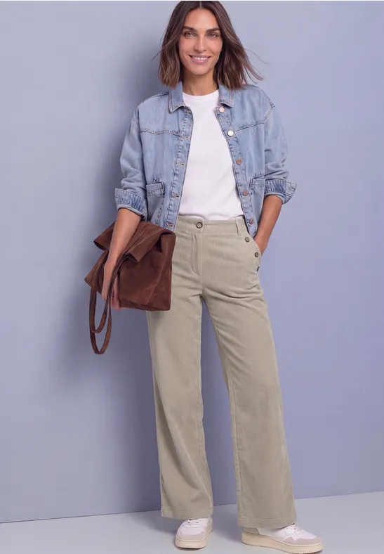 Eine Frau trägt Jeansjacke, weißes Shirt, weite beige Hose und hält eine braune Tasche.