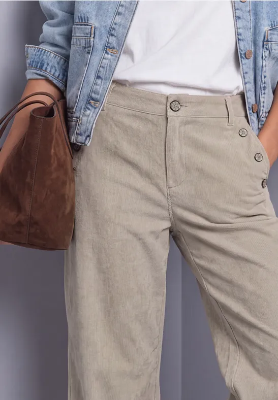 Frau trägt beige Cordhose mit seitlichen Knöpfen, weiße Bluse, Jeansjacke und braune Wildledertasche.