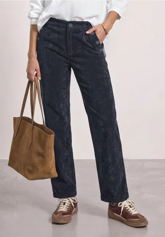 Gerade geschnittene, dunkelblaue Cordhose, weiße Oberteilkante sichtbar, braune Wildledertasche und Sneaker.