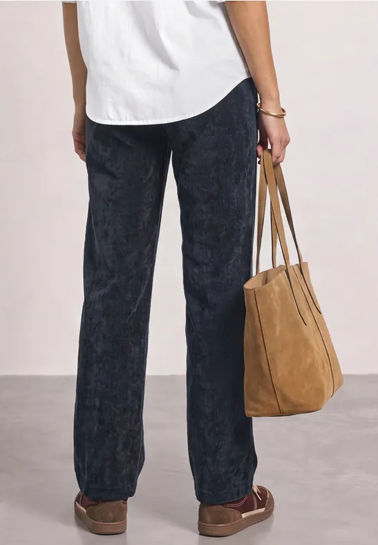 Rückansicht einer Person in schwarzen Cordhose, weißem Oberteil und braunen Loafers mit einer beigen Wildledertasche.