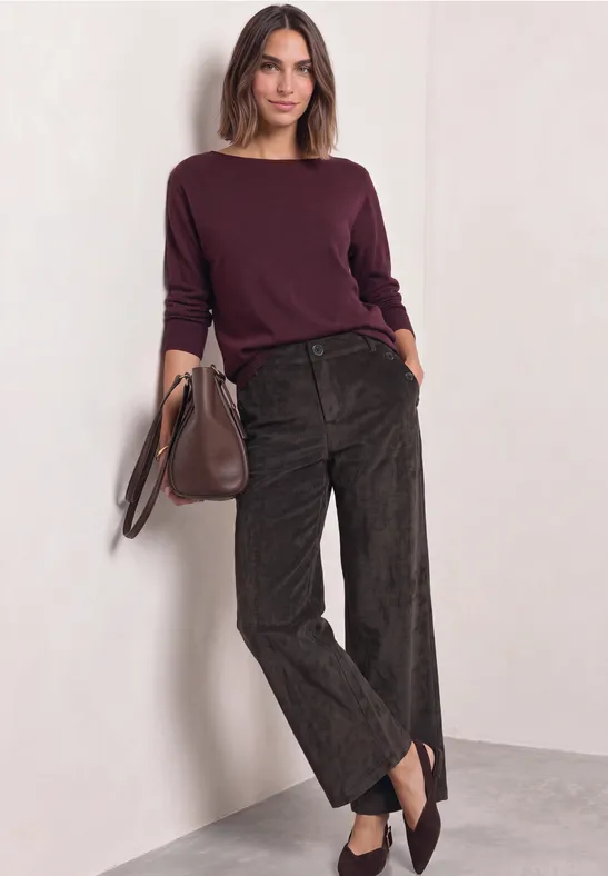 Frau lehnt an Wand, trägt bordeauxfarbenen Pullover, weite dunkelbraune Hosen und hält eine Tasche.
