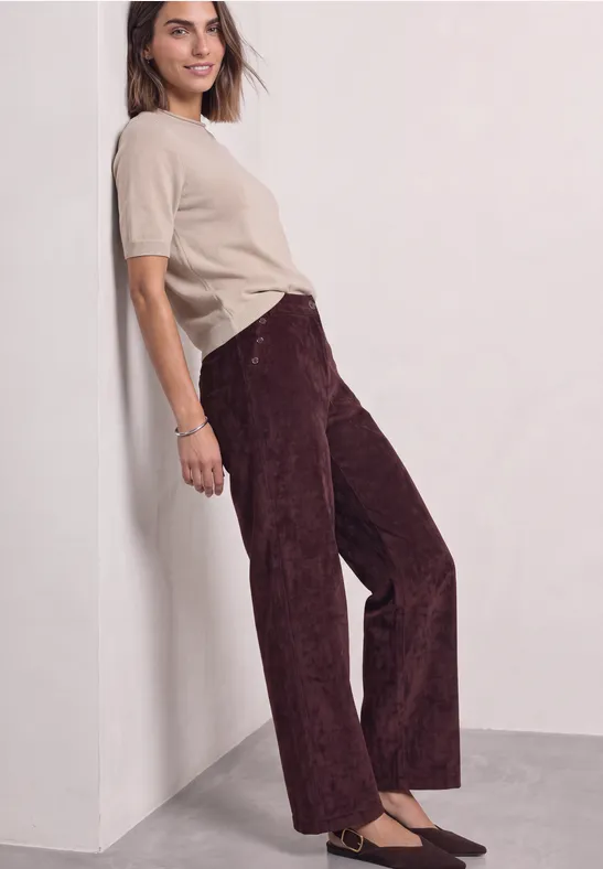Frau lehnt an Wand, trägt beige T-Shirt und bordeauxrote, weit geschnittene Cordhose.