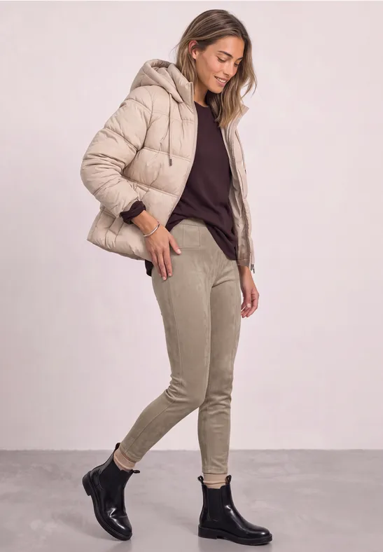 Frau in beiger Steppjacke, dunklem Pullover, hellen Hosen und schwarzen Stiefeletten, stehend vor hellem Hintergrund.